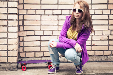 Blond teenage girl in a sunglasses with skateboard sits