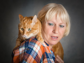 Beautiful aged woman hugging her red cat on grey background.
