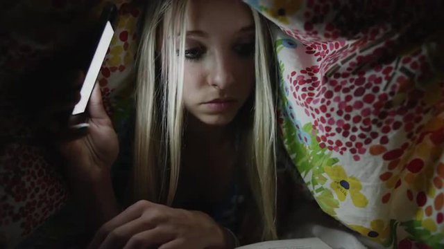 Close Up Of Teenage Girl Reading Book With Cell Phone Flashlight Under Blanket At Night / Sandy, Utah, United States