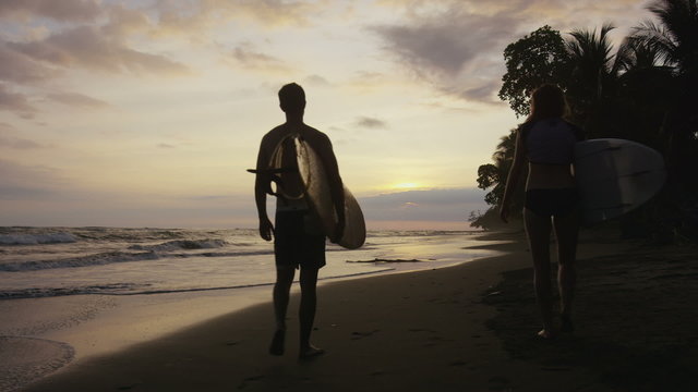 Wide shot of couple carrying surfboards on beach at sunset / Esterillos, Puntarenas, Costa Rica
