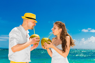 Happy bride and groom drink coconut water on a tropical beach. 