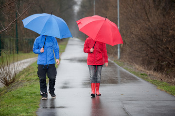 Regenspaziergang eines Paares durch einen Park in roter und blauer Regenkleidung mit Schirm 