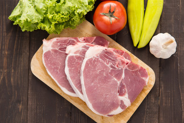 Raw pork on cutting board and vegetables on wooden background