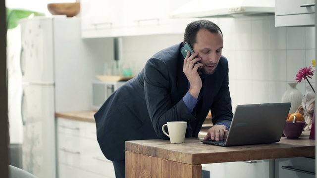 Young businessman with cellphone and laptop in kitchen at home
