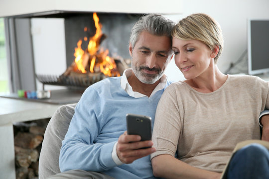 Middle-aged Couple Using Smartphone At Home By Fireplace