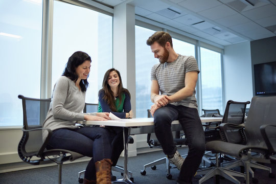 Three colleagues at a meeting, two women and a man seated on edge of the table. Looking over proofs of printed pages and designs. A creative environment.