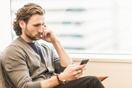 A bearded man sitting frowning and checking his phone.