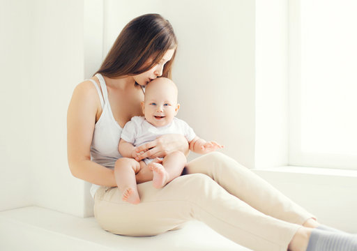 Lovely young mom kissing her baby at home in light room