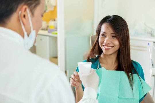 Smiling asian woman is taking a glass of water from her dentist