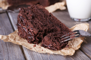 Sliced tasty chocolate cake on sheet of parchment and glass of milk on wooden table background