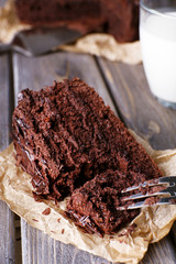 Sliced tasty chocolate cake on sheet of parchment and glass of milk on wooden table background