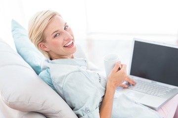 Obraz na płótnie Canvas Pretty blonde woman using her laptop and holding mug