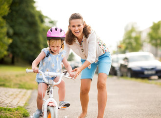 Happy mother pushes daughter on her bike as she learns to ride - Powered by Adobe