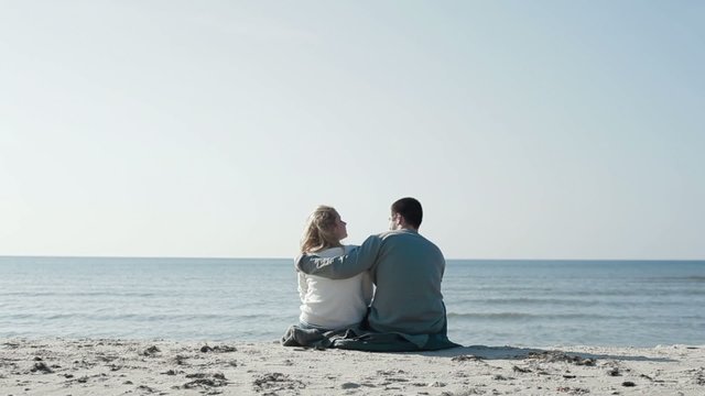 Slow motion of young happy couple in love sitting by the coast beach hugging looking at sea and enjoying romantic surroundings