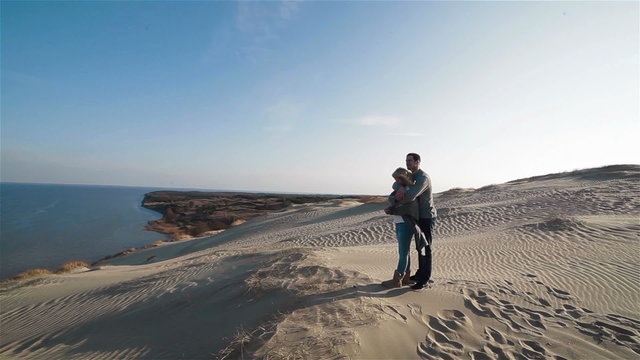 Beautiful flycam shot of young embracing couple standing on the top of enchanting natural sand dune hill and enjoying view to the sea