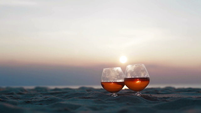 Close up of two glasses of brandy standing on the sand over incredible beach sunset background view. Male hand gallantly takes glasses away and then places back to the sand surface