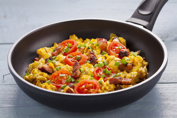 Scrambled eggs in a pan with bacon, onion and tomatoes sprinkled