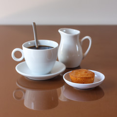 Cup of coffee, creamer jug and muffin on reflective table