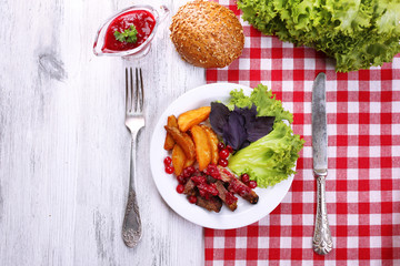 Beef with cranberry sauce, roasted potato slices, vegetables and bun on plate, on color wooden background