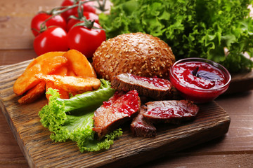 Beef with cranberry sauce, roasted potato slices and bun on cutting board, on wooden background