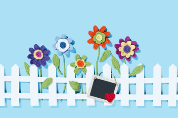 gehäkelte Blumen am Zaun mit Schild und Herz, blauer Hintergrund