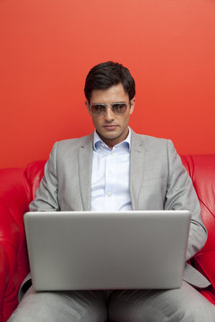Portrait of a businessman in a suit and sunglasses sitting on a red sofa using a laptop