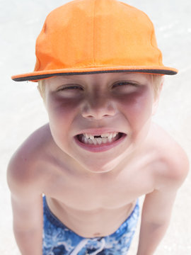 Close-up portrait of a shirtless boy in a baseball cap and shorts with a missing tooth standing on a beach pulling funny faces