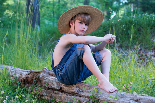 Young boy sitting on log wearing straw hat and blue overalls