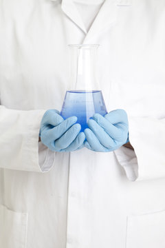 Close-up of a scientist in a laboratory coat and disposable gloves holding a conical flask filled with blue liquid