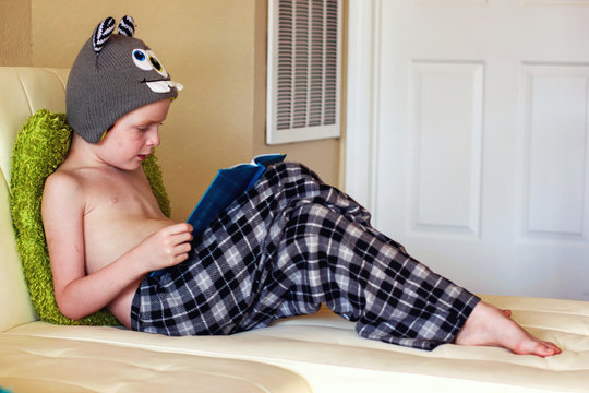 Side view of a shirtless boy in pyjama bottoms sitting on a sofa reading a book 
