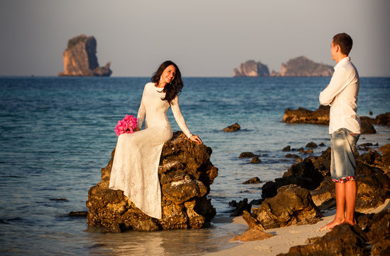 bride sits on rock groom stands on beach