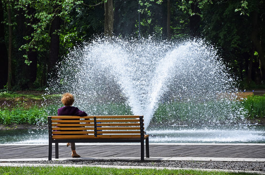 Old woman is resting on a park bench next to a fountain