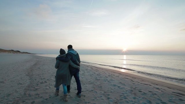 Young beautiful couple in love walking slowly side by side along coastline on the beach with wet sand by the sea, watching sunset and talking
