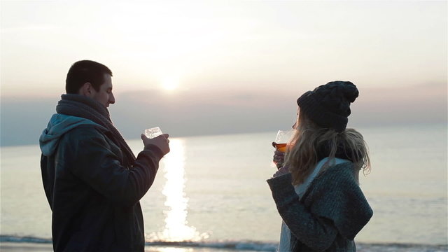 Young beautiful couple in love having warming drinks and enjoying the marvelous sunset sea view, discussing alcohol choice, smiling, holding hands and kissing 