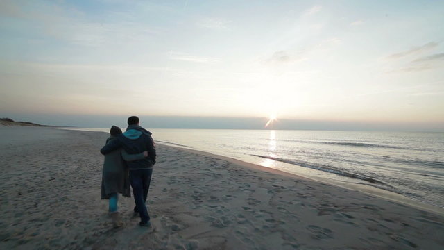 Slow motion of young couple in love walking slowly step by step side by side along coastline on the beach with wet sand by the sea, watching sunset and talking