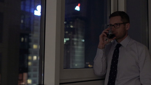Businessman standing next to the window and drinking wine at night
