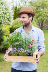 Happy smiling forty years old caucasian gardener holding wooden box full of flower seedlings in a pots. Spring garden as background.