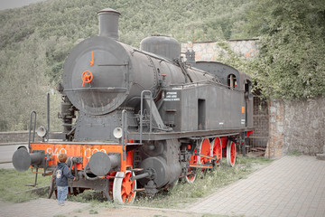 Fototapeta na wymiar Child observes fascinated from below the majestic front of an old locomotive FS 940 perfectly restored and exhibited to the public in a square of the Garfagnana, in Tuscany