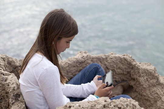 Niña sentada en roca junto al mar concentrada en su smartphone