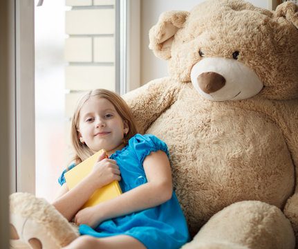 Little girl holding a book on windowsill with big toy