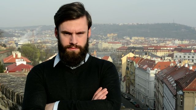 young handsome man with full-beard (hipster) looks to camera (confident look) - city in background
