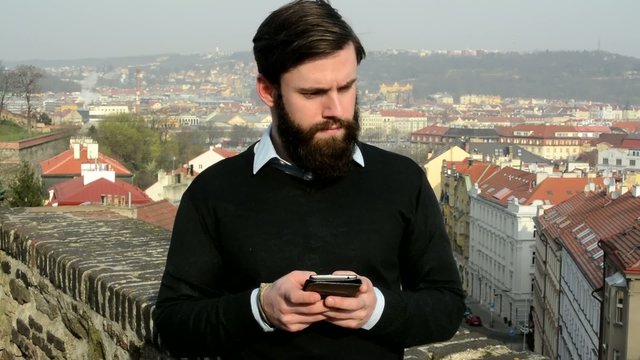 young handsome man with full-beard (hipster) works on mobile phone (serious face) - city in background