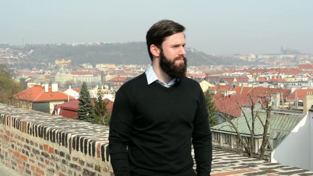 young handsome man with full-beard (hipster) walking in park and looks on the city - city in background