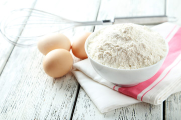 Bowl of wheat flour with eggs and whisk on white wooden backgrou