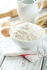 Bowl of wheat flour with eggs and whisk on white wooden backgrou