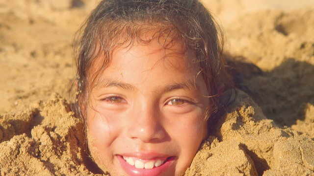 A young girl is buried in sand up to her neck. Smiling close up