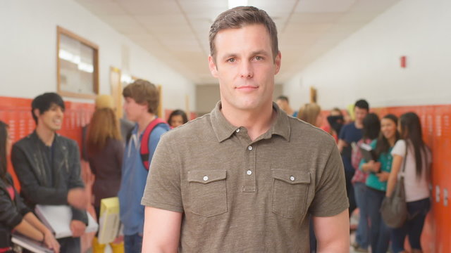 A male teacher stands in a busy hallway with students and smiles