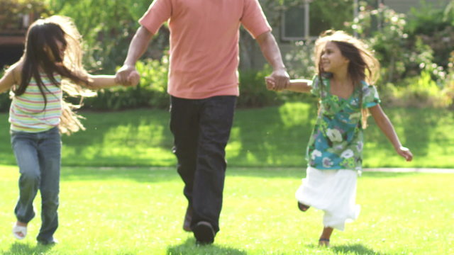 A father runs through an open green field with his two daughters on a sunny day