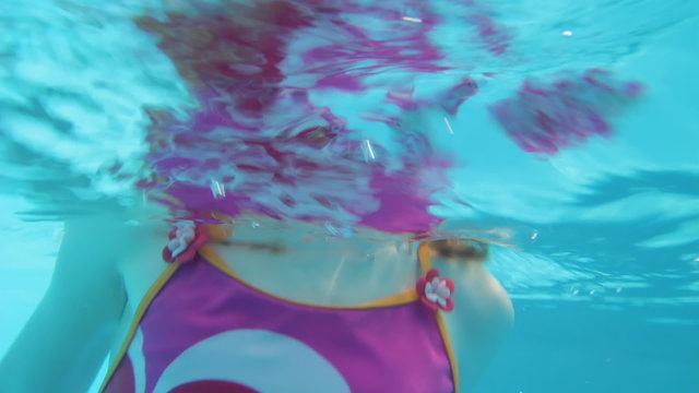 A young girl wearing goggles dives underwater and waves at the camera