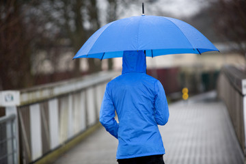 Ein Mann in blauer Regenkleidung und Schirm geht im Regen über eine Brücke